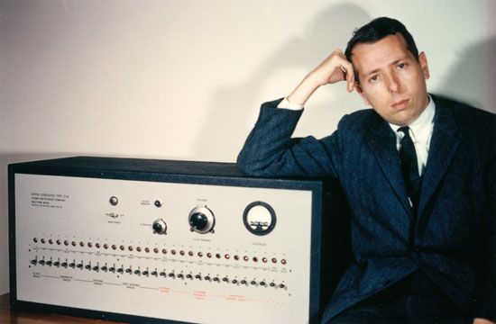 Milgram leans on an official looking old-fashioned piece of electrical equipment, with a long line of switches over some labels, and a few dials scattered about. The latter quarter of the labels are coloured red.