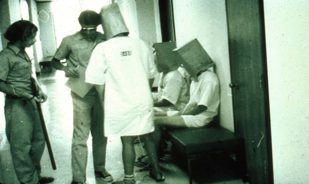 A still from the prison experiment. Prisoners in badly-fitting gowns have brown paper bags over their heads and sit or stand meekly before 70's style guards complete with long hair and aviator sunglasses, who observe with batons in hand.